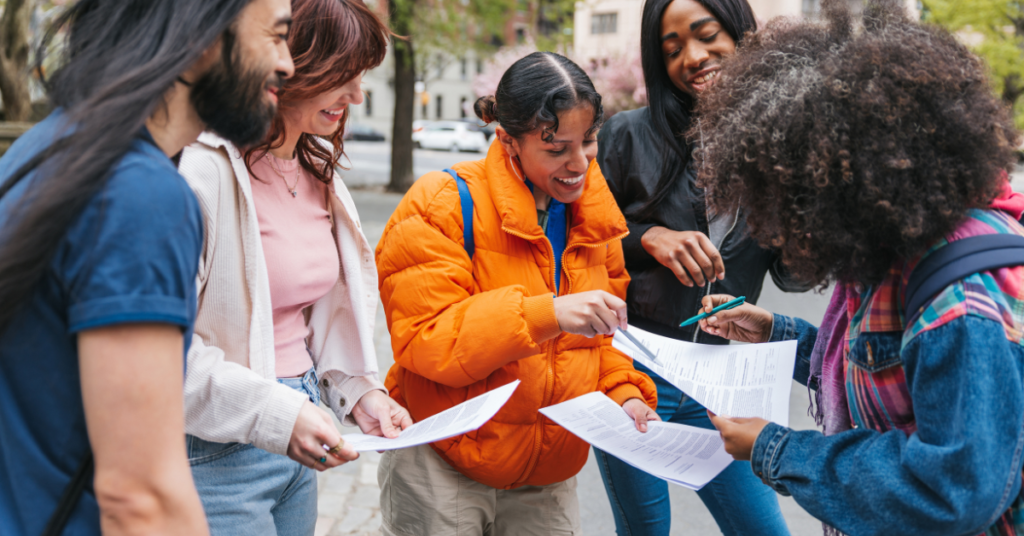 A diverse group of five individuals are gathered outdoors, engrossed in a lively discussion. They are holding and reviewing papers, with one woman in an orange jacket taking notes using a pen. The group appears cheerful and engaged, sharing a moment of collaboration and camaraderie amidst a city backdrop with trees in bloom.